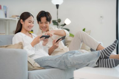 Genç çift, birlikte akıllı telefonlarıyla oyun oynamaktan zevk alıyor. Adam heyecan gösteriyor, kadın ise aydınlık, modern bir oturma odasında konsantre oluyor.