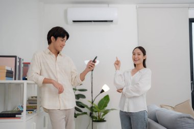 Mutlu genç çift, klima ünitesini açık, minimalist bir oturma odasında uzaktan kumandayla etkinleştiriyor. Birlikte rahat ve serin bir iklimin tadını çıkarıyorlar.