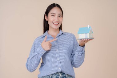 Mavi çizgili gömlekli neşeli genç kadın kameraya ev sahipliği, emlak yatırımı veya mimari konsepti simgeleyen küçük bir ev modelini sunuyor.