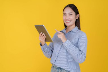 Çizgili gömlekli gülümseyen Asyalı iş kadını dijital tabletle poz veriyor, ekranı işaret ediyor, fotokopi alanı olan canlı sarı bir arka planda izole edilmiş.