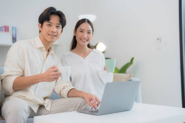 Neşeli genç çift bir dizüstü bilgisayarla birlikte oturur, parlak, modern bir ev ortamında kahvelerini yudumlar, rahatlama, ortaklık ve dijital yaşam tarzlarını aktarırlar.