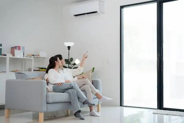 stock image Happy young couple lounges on a sofa in a bright, modern living room, using a remote control to adjust the air conditioning for enhanced home comfort