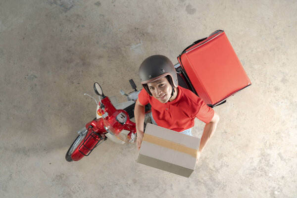 Top view of a delivery rider in red uniform with a helmet, preparing a cardboard package for dispatch next to a red motorcycle, on a concrete background