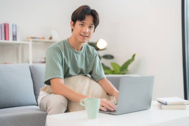 Kanepede oturan düşünceli genç adam iyi ışıklandırılmış oturma odasında dizüstü bilgisayarıyla çalışıyor. Odaklanmış ve biraz endişeli görünüyor. Yanında bir fincan ve kitaplarla.