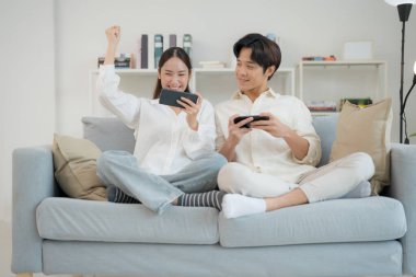 Genç çift, birlikte akıllı telefonlarıyla oyun oynamaktan zevk alıyor. Adam heyecan gösteriyor, kadın ise aydınlık, modern bir oturma odasında konsantre oluyor.