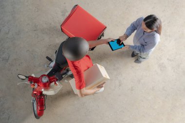 Kırmızı üniformalı bir teslimatçının gülümseyen bir kadın müşteriye bir paket teslim edişinin en üst görüntüsü. O da arka planda scooter ve teslimat kutusu olan mobil bir cihaz imzalıyor.