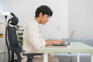 Odaklanmış genç bir adamın, parlak, modern bir ofis ortamında, bir fincan kahve ve rahat bir sandalyeyle uzaktan çalışmayı örnek alarak dizüstü bilgisayarda yazışının yan görüntüsü.