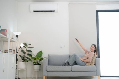 Kanepede uzanmış, elinde uzaktan kumandayla duvarlara monte edilmiş klimasını ayarlıyor, modern, parlak oturma odasındaki serin havayı takdir ediyor.
