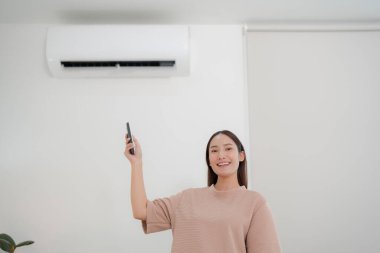 Parlak bir oturma odasında, rahat ve modern bir yaşam tarzının keyfini çıkararak, uzaktan kumandayla klima sıcaklığını ayarlayan, gündelik bir gömlekle gülümseyen genç bir kadın.