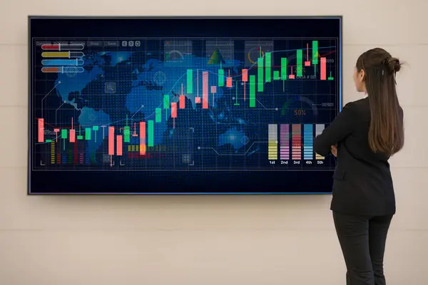 stock image Professional businesswoman stands in front of a large digital screen displaying colorful stock market charts and data for financial analysis and strategic planning in a corporate environment