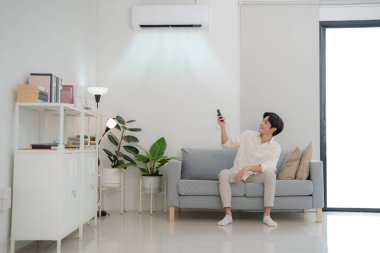 Genç adam bir koltukta oturuyor, klimayı uzaktan kumandayla kontrol ediyor, iyi ışıklandırılmış ve modern bir oturma odasında, konfor ve teknoloji dolu bir yaşam tarzı sergiliyor.