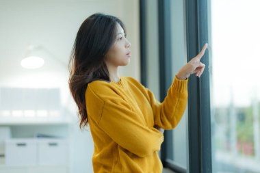 Canlı sarı kazaklı düşünceli genç bir kadın pencereden dışarı bakar, nazikçe cama dokunur, parlak, modern bir odada düşüncelere dalmıştır.