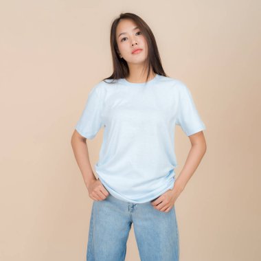 Genç Asyalı bir kadın tarafsız bir bej arka plana karşı duruyor, üzerinde klasik mavi bir kot pantolon bulunan düz bir mavi tişört var, basit ama modaya uygun bir tarzı temsil ediyor.