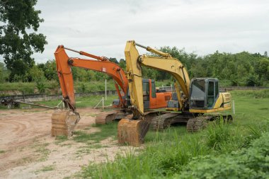 İki güçlü kazıcı yemyeşil çimlere park etmiş, işçiler için hazır. Ağır makineler, endüstriyel yapıları gösteren doğal manzarayla çelişiyor.