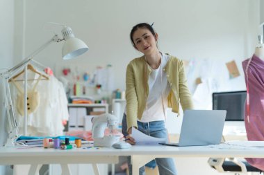 Genç moda tasarımcısı parlak stüdyosunda çalışıyor, kumaşı kesmek için işaretliyor. Etrafı mankenler, giysiler ve dikiş makinesiyle çevrili.
