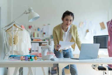 Genç moda tasarımcısı parlak stüdyosunda çalışıyor, kumaşı kesmek için işaretliyor. Etrafı mankenler, giysiler ve dikiş makinesiyle çevrili.