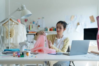 Genç moda tasarımcısı parlak stüdyosunda çalışıyor, kumaşı kesmek için işaretliyor. Etrafı mankenler, giysiler ve dikiş makinesiyle çevrili.