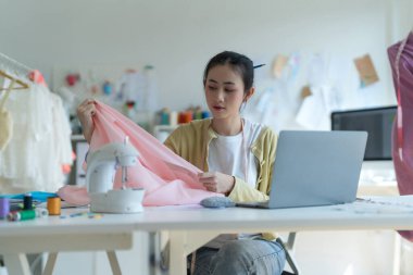 Genç moda tasarımcısı parlak stüdyosunda çalışıyor, kumaşı kesmek için işaretliyor. Etrafı mankenler, giysiler ve dikiş makinesiyle çevrili.