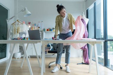 Genç moda tasarımcısı parlak stüdyosunda çalışıyor, kumaşı kesmek için işaretliyor. Etrafı mankenler, giysiler ve dikiş makinesiyle çevrili.