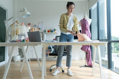 Genç moda tasarımcısı parlak stüdyosunda çalışıyor, kumaşı kesmek için işaretliyor. Etrafı mankenler, giysiler ve dikiş makinesiyle çevrili.