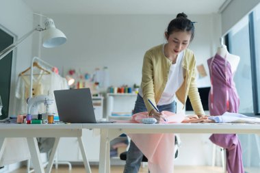 Genç moda tasarımcısı parlak stüdyosunda çalışıyor, kumaşı kesmek için işaretliyor. Etrafı mankenler, giysiler ve dikiş makinesiyle çevrili.