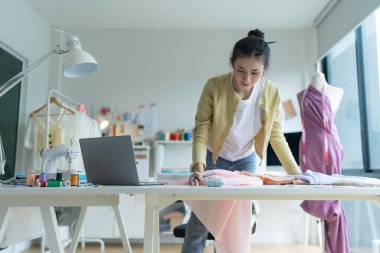 Genç moda tasarımcısı parlak stüdyosunda çalışıyor, kumaşı kesmek için işaretliyor. Etrafı mankenler, giysiler ve dikiş makinesiyle çevrili.