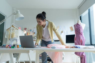 Genç moda tasarımcısı parlak stüdyosunda çalışıyor, kumaşı kesmek için işaretliyor. Etrafı mankenler, giysiler ve dikiş makinesiyle çevrili.