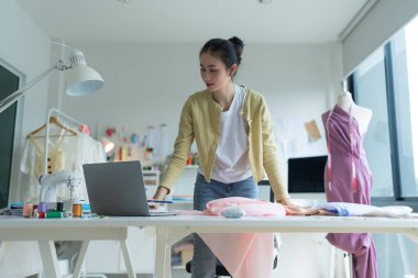 Genç moda tasarımcısı parlak stüdyosunda çalışıyor, kumaşı kesmek için işaretliyor. Etrafı mankenler, giysiler ve dikiş makinesiyle çevrili.