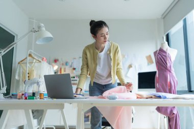 Genç moda tasarımcısı parlak stüdyosunda çalışıyor, kumaşı kesmek için işaretliyor. Etrafı mankenler, giysiler ve dikiş makinesiyle çevrili.