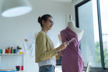 Çekici genç moda tasarımcısı elinde tabletle stüdyoda duruyor. Tasarladığı kıyafetlerle mankenin yanında..