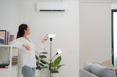 Genç bir kadın klimayı ayarlamak için uzaktan kumanda kullanıyor. Minimalist dekorlu, modern bir oturma odasında.