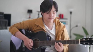 Genç adam rahat bir şekilde rahat bir oturma odasında kanepede oturur, akustik gitar çalar. Atmosfer, müzik için rahatlama ve tutku yayıyor. Yeteneğini ve yaratıcılığını gösteriyor.