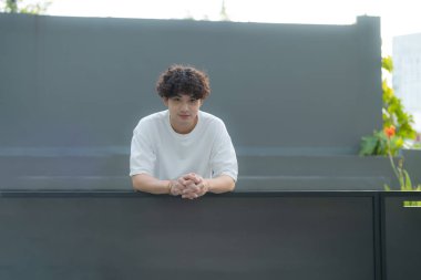 Kıvırcık saçlı ve beyaz tişörtlü genç bir adam, çağdaş bir açık alanda gri bir duvara yaslanmış, sıradan ama şık bir hava yayıyor.