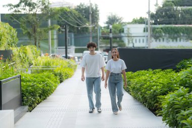 Beyaz t-shirtler ve kot pantolon giyen mutlu genç Asyalı çift modern bir şehirde yürüyorlar, birlikte keyifli bir gezinti yapıyorlar.