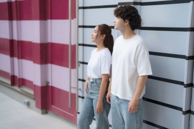 Beyaz t-shirt ve kot giyen genç bir adam ve kadın renkli bir duvara yaslanmış, uzaklığa bakıyorlar.