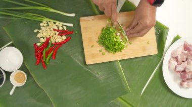 Şef ahşap bir kesim tahtasının üzerinde yeşil soğan doğruyor, domuz eti, sarımsak, biber ve muz yaprakları üzerinde çeşitli baharatlarla lezzetli bir yemek hazırlıyor.