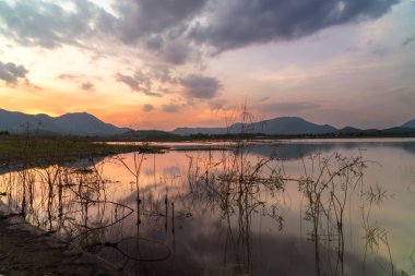 Sakin göl, arka planda görkemli dağlarla çevrili canlı günbatımı renklerini ve ön planda yemyeşil otları yansıtıyor. Huzurlu ve resimsel bir manzara yaratıyor.