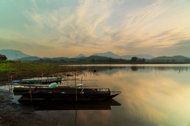 Sakin bir gölün yüzeyine yansıyan canlı günbatımı renkleri, arka planda dağların siluetleri ve sakin manzarayı tamamlayan bulutlu bir gökyüzü.