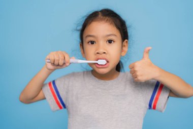 Asyalı bir kız diş fırçasıyla dişlerini fırçalıyor, canlı bir mavi zemine karşı diş hijyeninin önemini vurguluyor, mutluluk saçıyor ve sağlıklı alışkanlıkları teşvik ediyor.