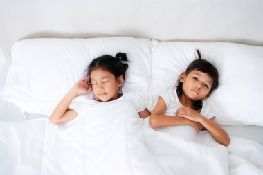 İki küçük Asyalı kız huzur içinde uyuyorlar ve beyaz battaniyenin altında el ele tutuşuyorlar. Dinlenme ve bağlanma anlarının tadını çıkarıyorlar.