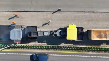 Otoyoldaki bir inşaat alanının havadan çekilmiş görüntüleri. İçinde büyük bir asfalt döşeme makinesi ve işçiler var. Sitede kısmen tamamlanmış bir köprü veya yol bölümü gösteriliyor.