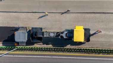 Otoyoldaki bir inşaat alanının havadan görünüşü, büyük bir asfalt döşeme makinesinin ve işçilerin yer aldığı. Site kısmen tamamlanmış bir yol parçasını gösteriyor.