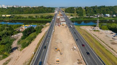 Büyük bir vinç, inşaat malzemeleri ve işçilerin yer aldığı otoyoldaki bir inşaat alanının havadan görüntüsü. Sitede kısmen tamamlanmış bir köprü veya yol bölümü yer alıyor..