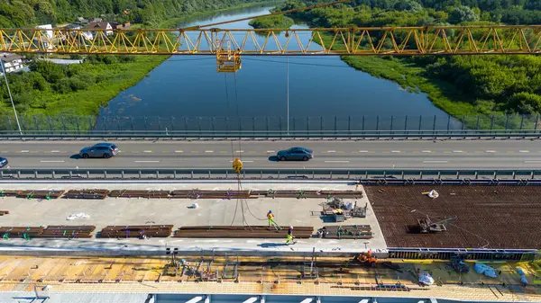Büyük bir vinç, inşaat malzemeleri ve işçilerin yer aldığı otoyoldaki bir inşaat alanının havadan görüntüsü. Sitede kısmen tamamlanmış bir köprü veya yol bölümü yer alıyor..