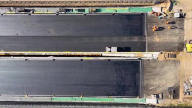 Otoyoldaki bir inşaat alanının havadan çekilmiş görüntüleri. İçinde büyük bir asfalt döşeme makinesi ve işçiler var. Sitede kısmen tamamlanmış bir köprü veya yol bölümü gösteriliyor.