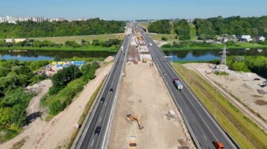 Büyük bir vinç, inşaat malzemeleri ve işçilerin yer aldığı bir otoyoldaki inşaat alanının hava görüntüleri. Sitede kısmen tamamlanmış bir köprü veya yol bölümü yer alıyor..