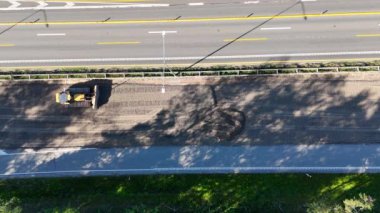 Yol kenarındaki bir yol şantiyesinin havadan çekilmiş görüntüsü, yeni derecelendirilmiş bir yüzeyde büyük sarı bir buldozer bulunuyor. Bölge yakındaki ağaçların gölgelerini ve şerit işaretli yolu gösteriyor..