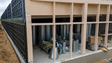 Büyük pencereli ahşap bir yapıya sahip inşaat alanı. İçinde çeşitli metal kanallar ve ekipmanlar, kutular ve inşaat malzemeleriyle birlikte görülebilir durumda. Dış yüzeyde iskele ve tamamlanmamış zemin görünüyor..