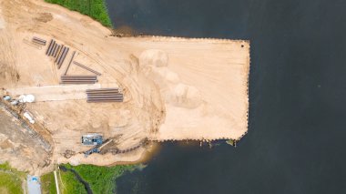 Nehir kıyısındaki bir inşaat alanının havadan görünüşü, kum yığınları ve ahşap yapılarla dolu kumlu bir alan. Çevredeki yeşillikler ve binalar görülebilir..
