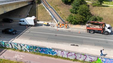 Yol kenarındaki bir inşaat alanının havadan görüntüsü. İşçiler bir kamyonet ve beyaz bir teslimat aracının yanında görülmüş..